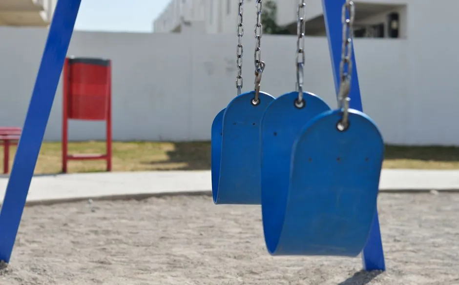 Ein leerer Spielplatz im Freien mit einer Reihe blauer Schaukelsitze, still an Metallketten hängend, eine rote Bank unscharf im Hintergrund, keine Personen, kein lesbarer Text