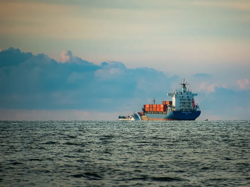 Ein großes, mit Containern beladenes Containerschiff fährt bei Dämmerung auf offener See, daneben ein kleines Lotsenboot, keine Gesichter oder lesbaren Texte sichtbar