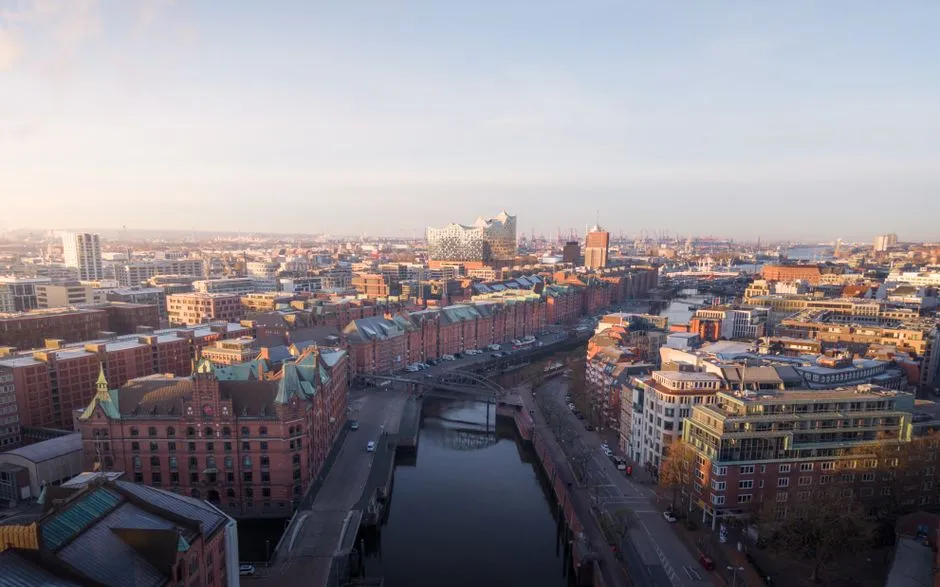 Vista aérea del Speicherstadt de Hamburgo a la luz del día, hileras de almacenes de ladrillo rojo con tejados de cobre verde bordeando una red de canales y puentes, con el tejado de cristal ondulado de la Elbphilharmonie visible al fondo, sin personas ni señalización legible
