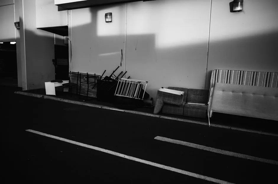 A black and white photo of an old sofa, wooden chairs, a folded mattress, and stacked chairs set against a building wall at a curbside, no people or readable text visible