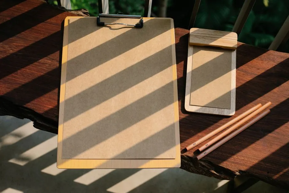 Ein leeres Holz-Klemmbrett, ein kleiner Holznotizblock und drei angespitzte Bleistifte auf einem sonnenbeschienenen Holztisch, keine Gesichter, Logos oder lesbaren Texte sichtbar