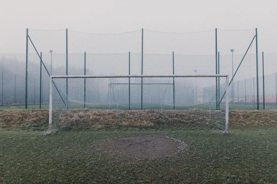 Leeres Fussballtor mit vollstaendigem Netz auf einem frostigen, nebligen Platz im Morgengrauen, hoher Zaun im Hintergrund, keine Spielerinnen, Spieler, Logos oder lesbare Beschilderung sichtbar
