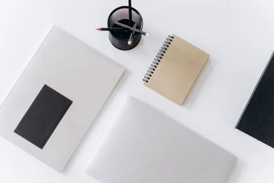Un portátil cerrado, un cuaderno de espiral, lápices en un pequeño soporte, y una carpeta dispuestos sobre un escritorio blanco liso, sin rostros ni texto legible visible