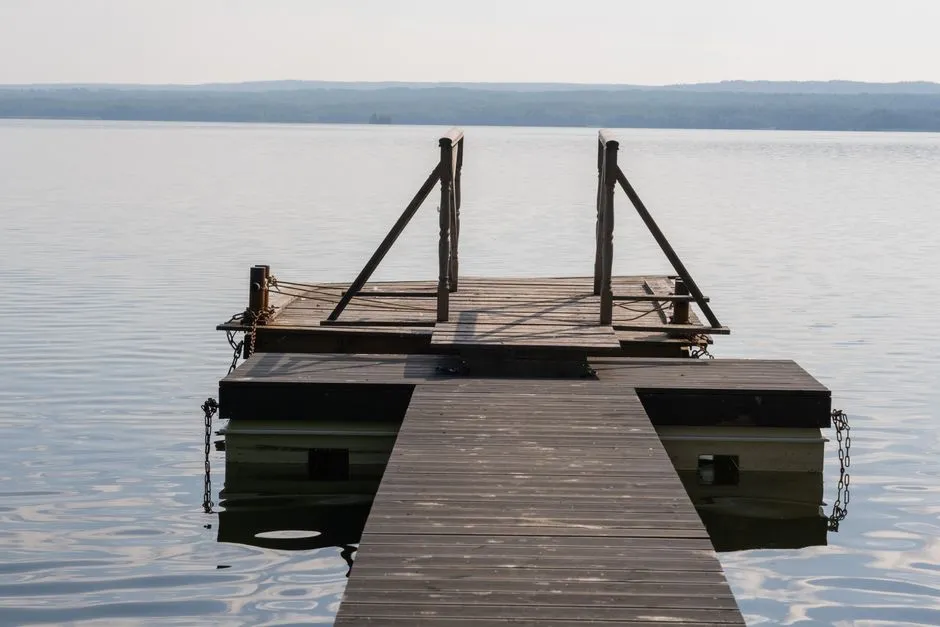 Ein leerer Holzsteg erstreckt sich über die ruhige Oberfläche eines Sees, baumgesäumtes Ufer in der Ferne, keine Menschen sichtbar