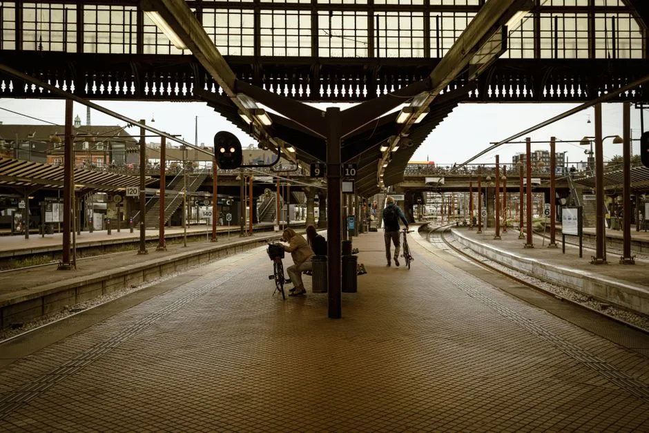 Ein überdachter Bahnsteig mit kunstvollem Glasdach darüber und in die Ferne führenden Gleisen, zwei Personen weit hinten auf dem Bahnsteig stehen jeweils neben einem Fahrrad, aus der Distanz fotografiert, keine Gesichter erkennbar, keine lesbaren Beschilderungen oder Logos