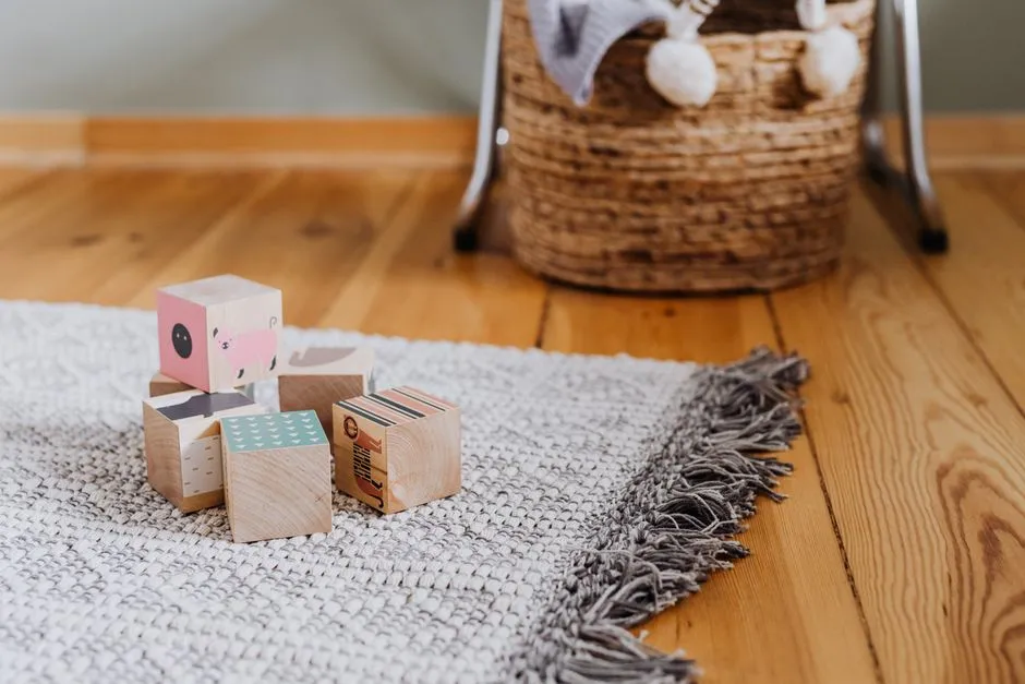 Ein kleiner Haufen hölzerner Bauklötze mit einfach gemalten Tier- und Mustermotiven, verstreut auf einem gewebten grauen Teppich neben einem Weidenkorb, auf einem Holzboden