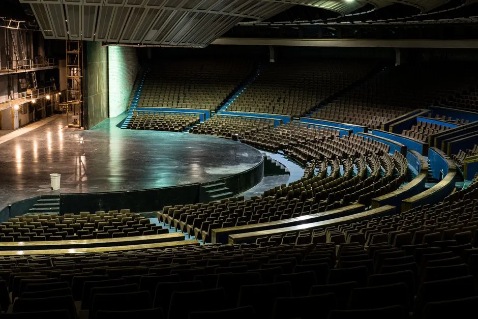 Wide view of an empty theater-in-the-round seen from above, curved rows of dark seats wrapping around a round stage, backstage scaffolding visible to one side, no people or readable signage visible
