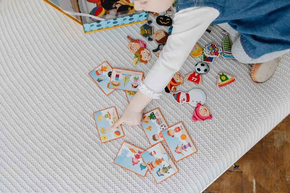 Die Hand eines Kindes zeigt auf eine Auswahl bunter Bildkarten, ausgebreitet auf einer gestrickten Decke, mit einem kleinen Stapel ähnlicher Karten daneben