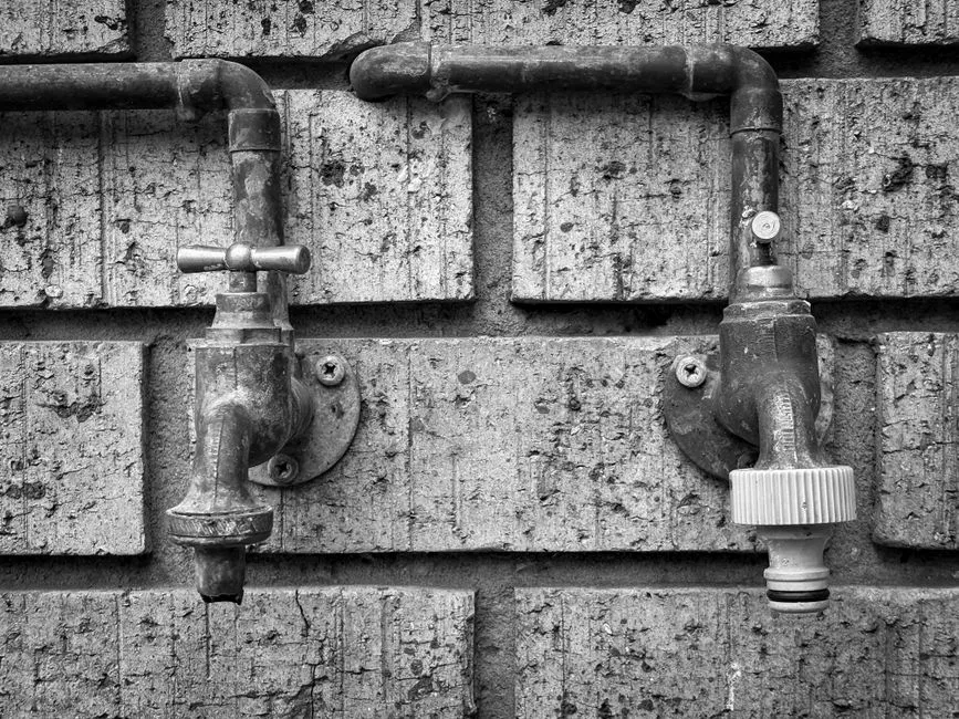 Two old metal outdoor water taps with round shut-off valves mounted on a brick wall, black and white, no people or readable text visible