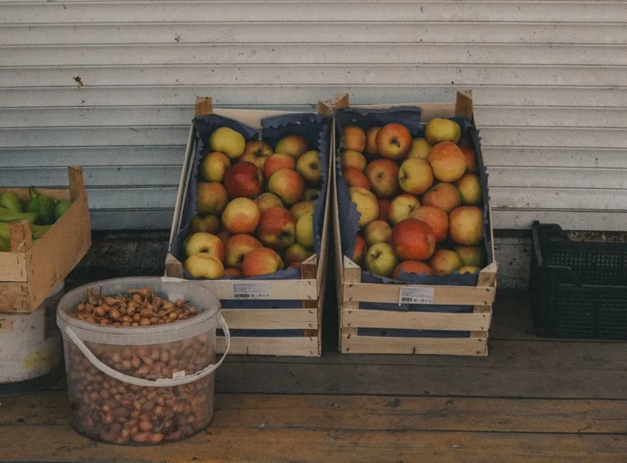 Cajas de madera con manzanas frescas y un cubo de chalotas expuestos afuera de un puesto de mercado, sin personas ni carteles visibles