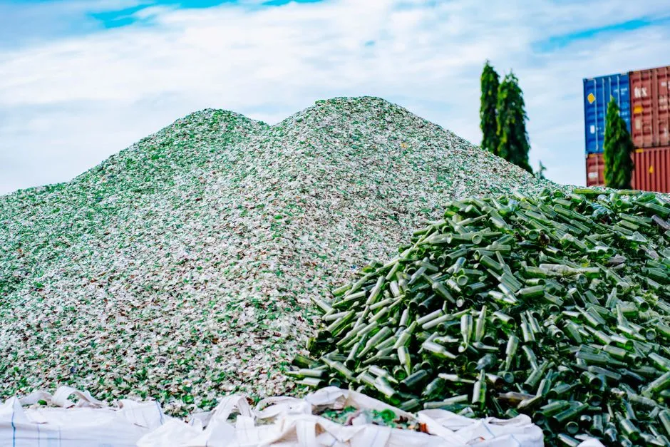 A large industrial pile of crushed green glass cullet and stripped green glass bottles at an outdoor recycling facility, no people or readable text visible