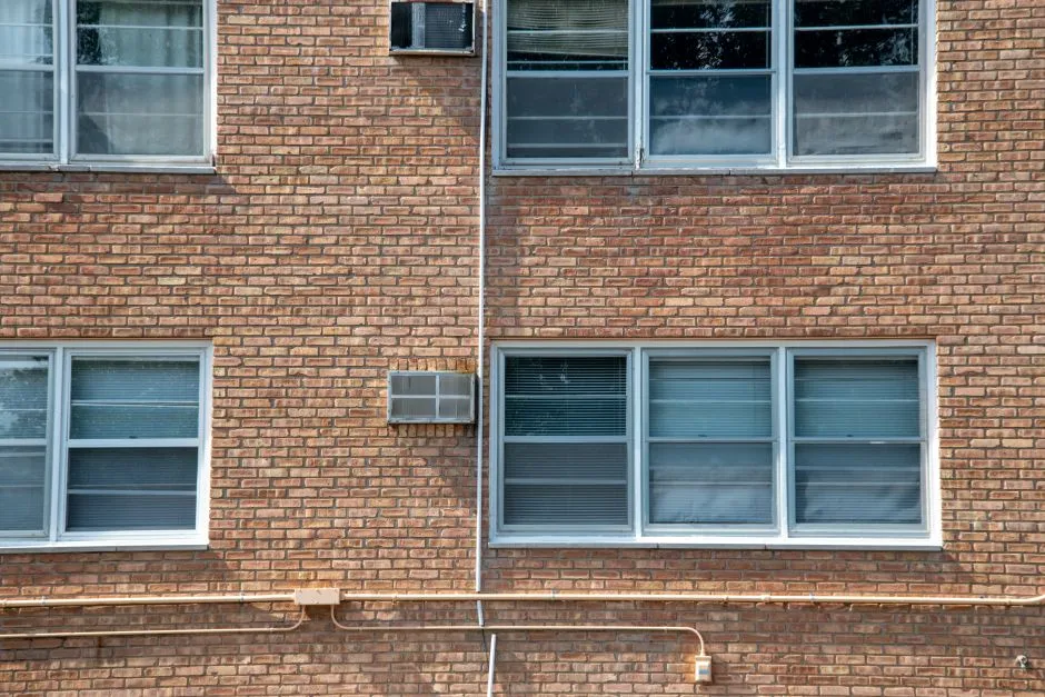 Eine Reihe von Fenstern einer Backstein-Wohnhausfassade, mit fensterseitig montierten Klimaanlagen, ohne erkennbaren Text oder Personen