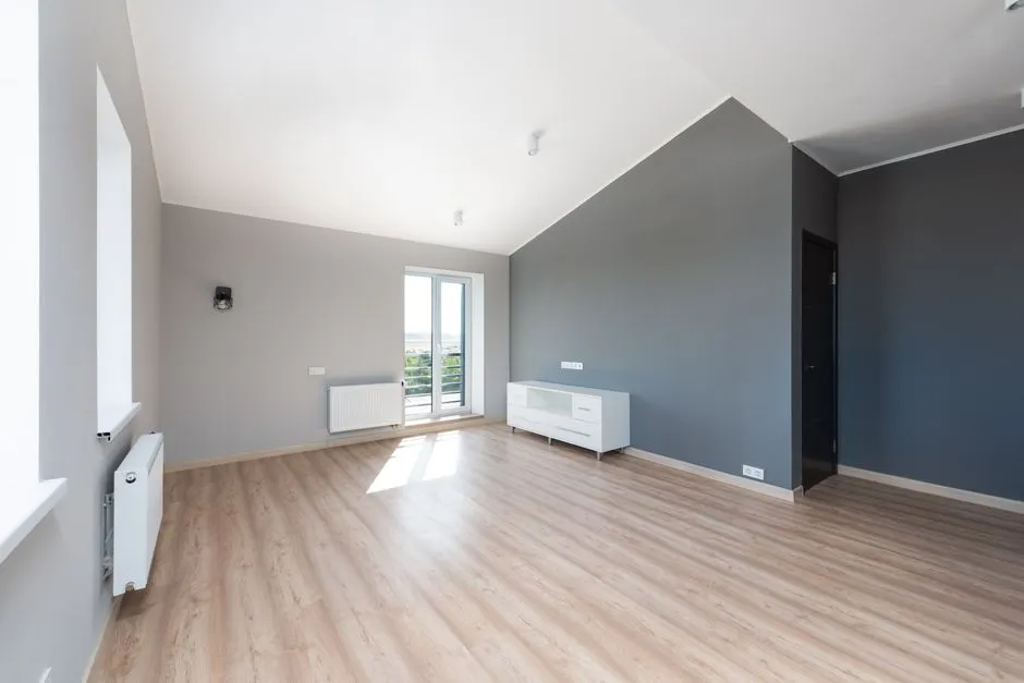 A clean, freshly renovated empty apartment room with light wood flooring, gray walls, a radiator, and a glass balcony door, with no furniture or people visible