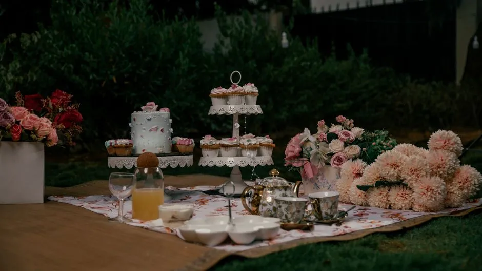 Ein eleganter Tee-Party-Tisch im Freien in der Abenddämmerung, gedeckt mit einer blumengeschmückten Torte, einer mehrstöckigen Cupcake-Etagere, Teetassen, einer Teekanne, sowie Rosen- und Dahlienstraußen, keine Menschen sichtbar