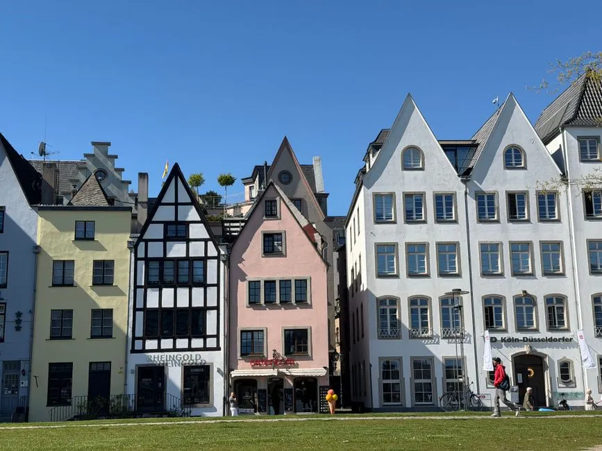 A row of colorful gabled historic houses in Cologne's Altstadt, painted yellow, black-and-white timber-frame, pink, and white, under a clear blue sky