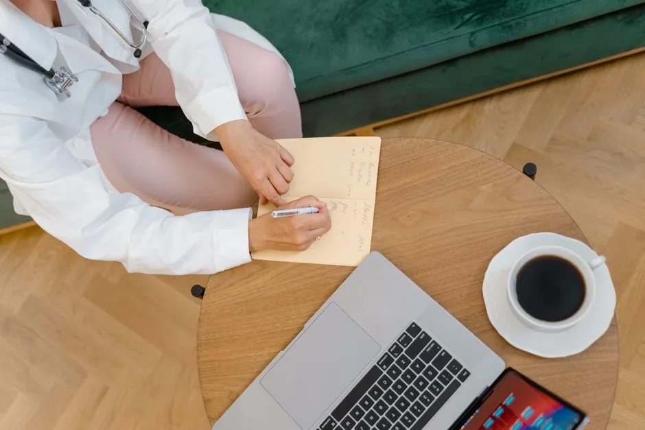 Eine Ärztin mit Stethoskop macht sich Notizen an einem Couchtisch neben einem Laptop und einer Tasse Kaffee