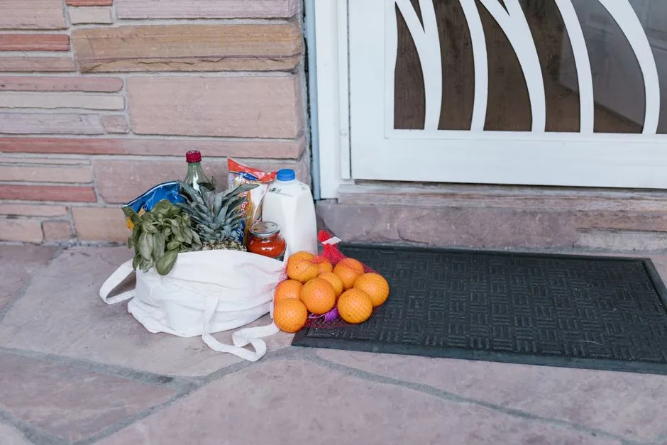 Eine Stofftasche voller frischer Lebensmittel, darunter eine Ananas, Basilikum, Milch und ein Netz Orangen, auf einer Türschwelle abgestellt