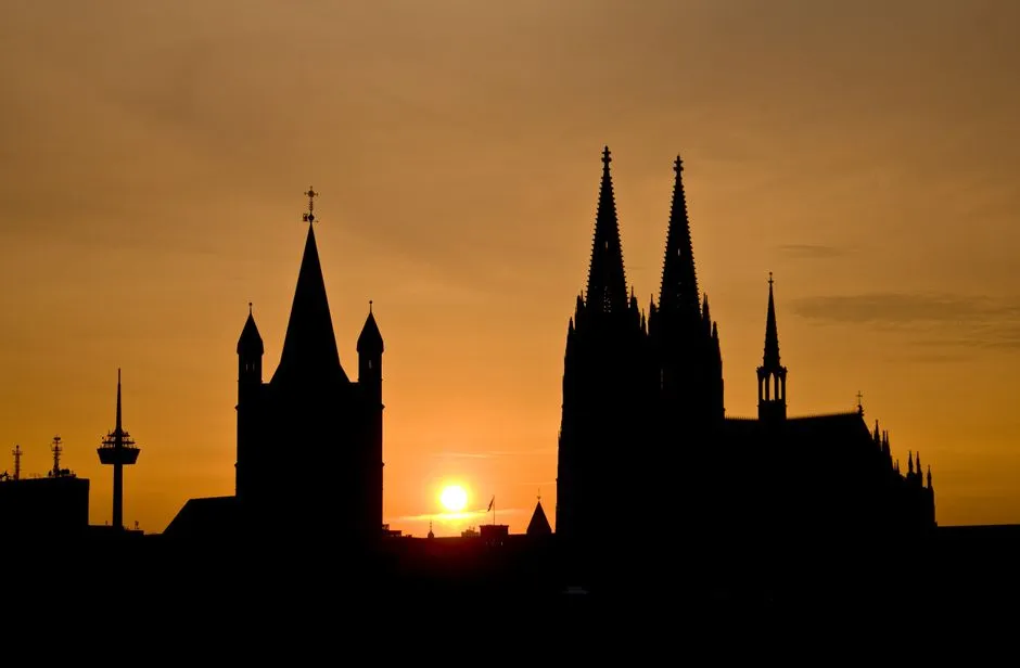 Silhouette der beiden gotischen Türme des Kölner Doms vor orangefarbenem Sonnenuntergangshimmel, mit dem Kölner Fernsehturm links im Bild