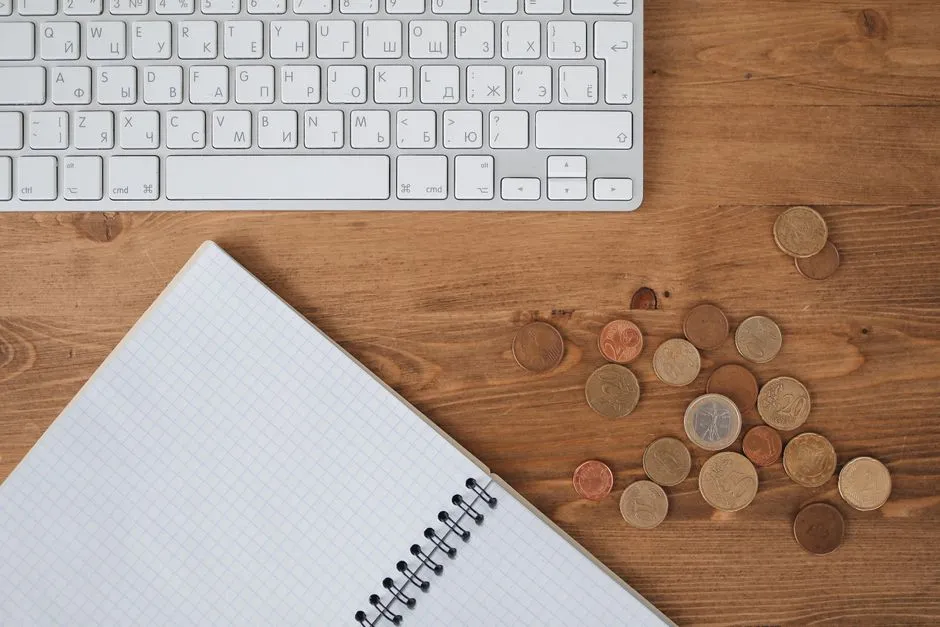 A computer keyboard, a spiral-bound notebook, and a small scattered pile of euro coins on a wooden desk