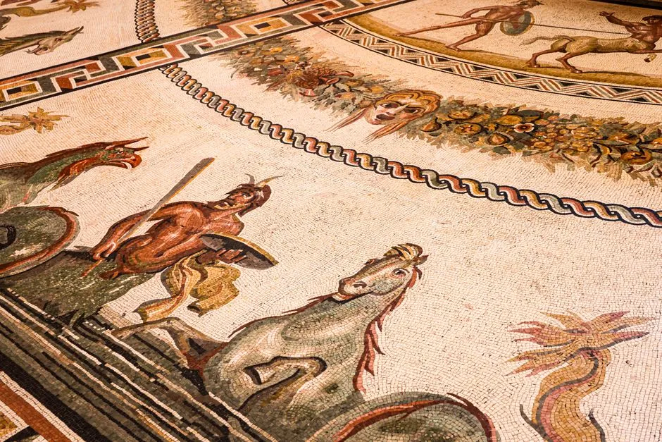 Nahaufnahme eines antiken römischen Bodenmosaiks mit fein detaillierten Figuren, Tieren und geometrischen Randmustern in Beige-, Rot- und Dunkeltönen