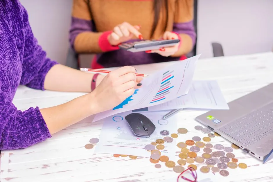 Two people reviewing a budget chart with a calculator, laptop, and loose coins on a table
