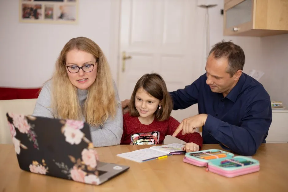 A parent filling out an official form online on a laptop at a kitchen table while a young child sits nearby