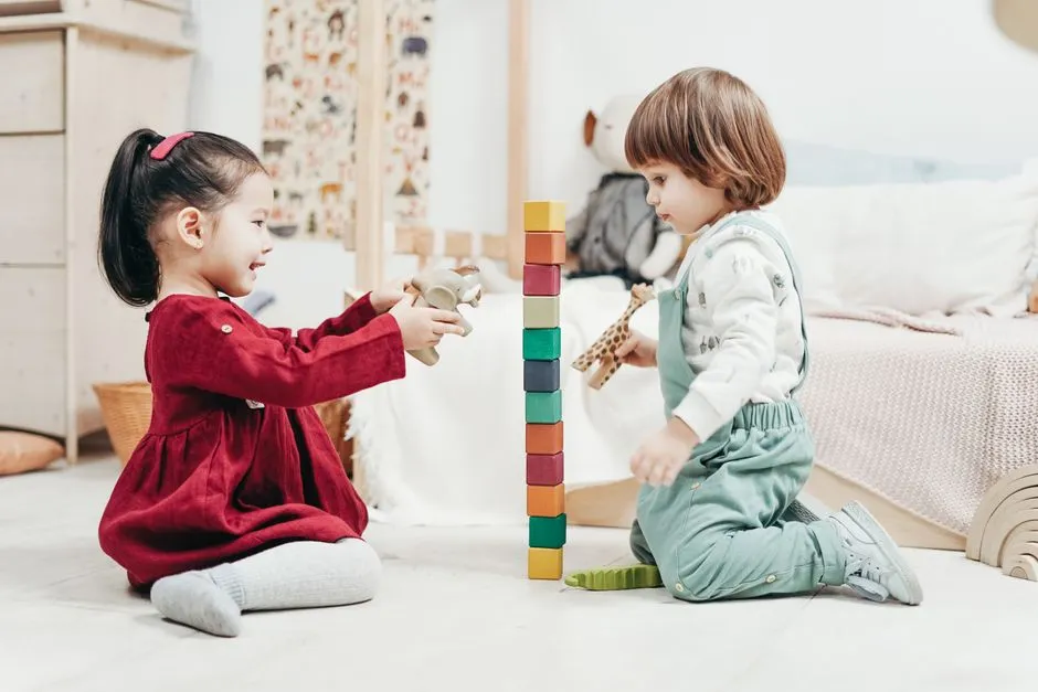 Two toddlers playing with a stack of colorful wooden building blocks on the floor of a bright playroom
